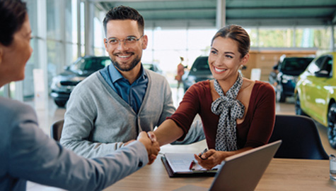 A man and a woman are shaking hands while sitting at a table with a laptop at a car dealership